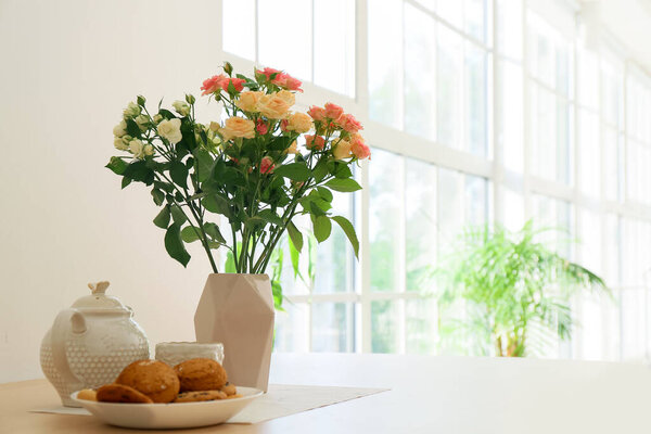 Ваза с красивыми розами, чайник и печенье на обеденном столе на кухне