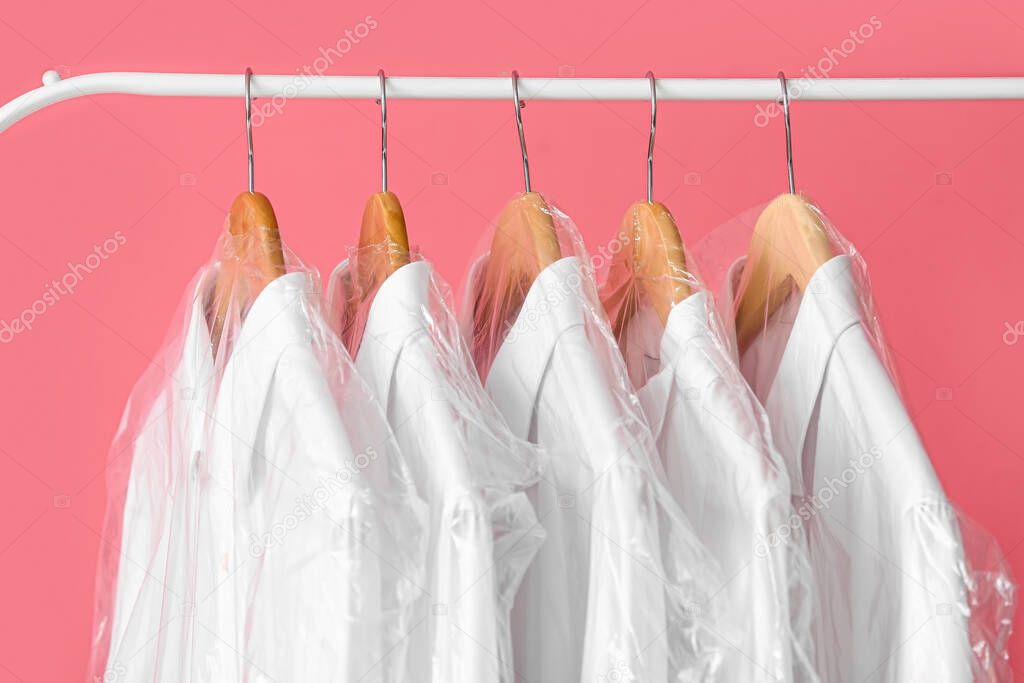 Rack with clean white shirts in plastic bags on pink background, closeup