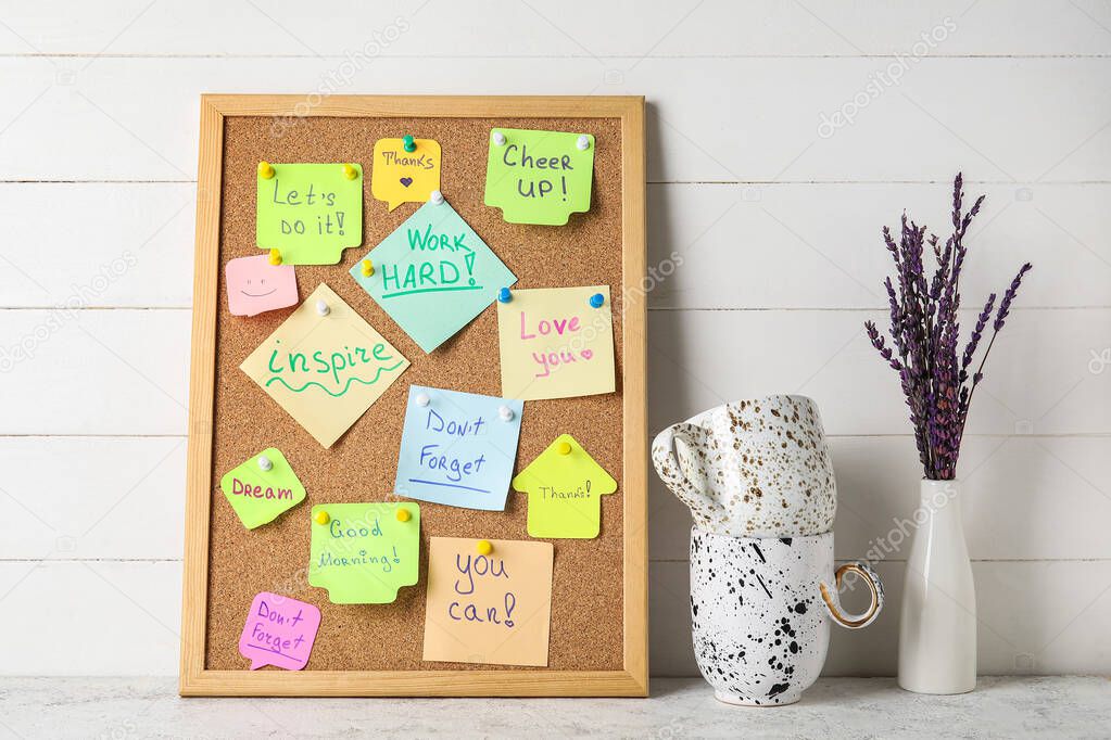 Board with different sticky notes, cups and vase on white wooden background