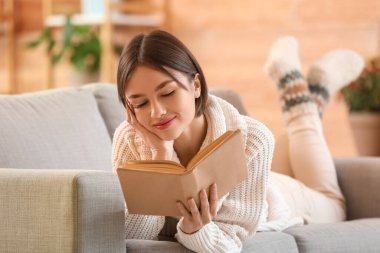 Evdeki kanepede kitap okuyan güzel genç bir kadın.