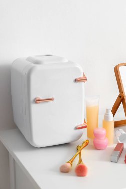 Işık duvarının yanındaki masada kozmetik ürünleri ve makyaj fırçaları olan küçük bir buzdolabı.