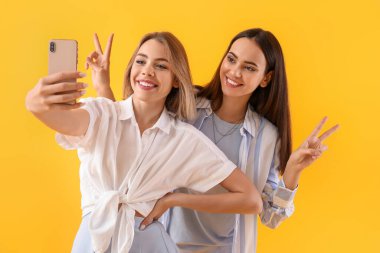 Cep telefonlu genç kadınlar sarı arka planda selfie çekiyorlar.