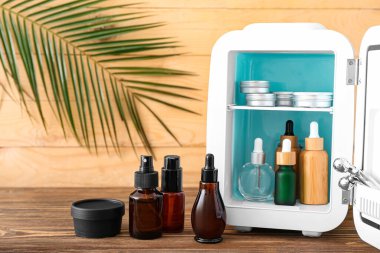 Kozmetik ürünleri ve ahşap masa üzerinde tropikal yapraklı küçük buzdolabı.
