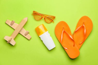 Tahta uçak, güneş gözlüğü, bir şişe güneş kremi ve yeşil arka planda parmak arası terlikler.