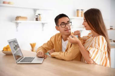 Genç çift mutfaktaki masada dizüstü bilgisayar kullanıyor.