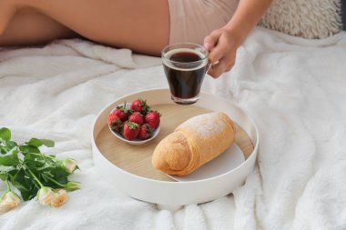 Genç bir kadın tepsiden kahve alıyor. Yatakta lezzetli bir kahvaltı, yakın plan.
