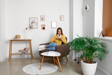 Kulaklığı ve sıcak içeceği olan güzel bir kadın evdeki yumuşak koltukta oturuyor.