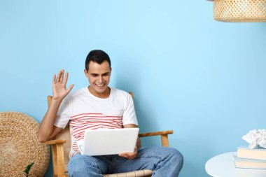 Genç adam mavi duvarın yanındaki tahta koltukta dizüstü bilgisayarla sohbet ediyor.