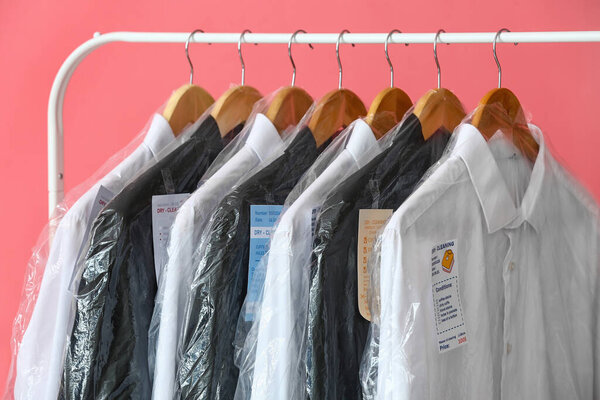 Rack with clean clothes in plastic bags on pink background