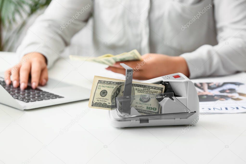 Female accountant working with laptop and currency counting machine on light table