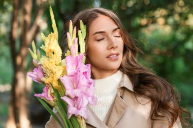 Kapalı gözlü ve bir buket Gladiolus çiçekli bir kadın parkta güneşli bir günün tadını çıkarıyor.