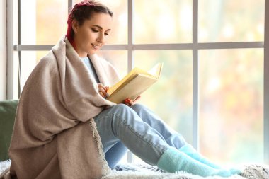 Ekose kumaşlı güzel bir kadın evde pencere eşiğinde kitap okuyor.