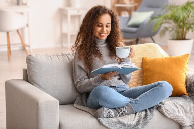 Evdeki kanepede çay fincanı okuyan genç Afro-Amerikalı kadın.