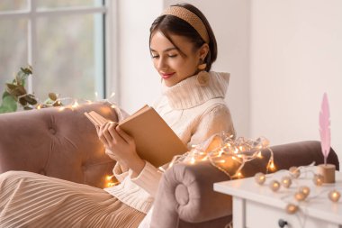 Evdeki kanepede kitap okuyan güzel genç bir kadın.