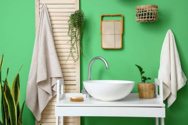 Banyoda yeşil duvarın yanında beyaz lavabosu, çiçekleri ve sabunu olan bir masa.