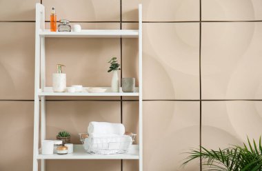 Bej duvarın yanında banyo aksesuarları olan raf ünitesi