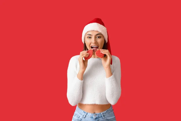 戴着圣诞礼帽的快乐年轻女人吃着红色背景的姜饼 — 图库照片