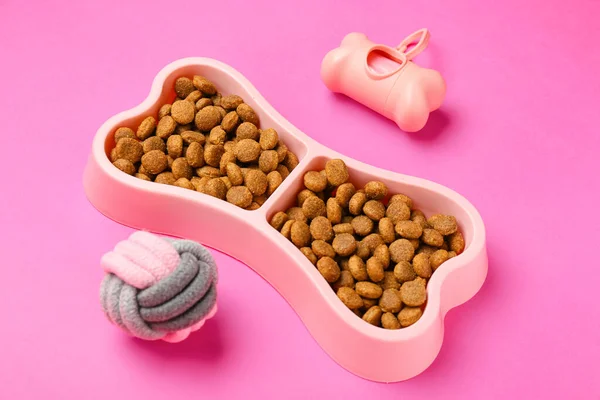 Bowl with dry pet food, toy ball and waste bags on color background