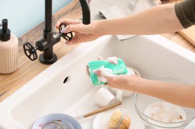 Temizlik süngeri olan kadın mutfaktaki musluğu açıyor.