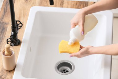 Ev hanımı mutfaktaki lavabonun üzerinden deterjanı süngerin üzerine döküyor.