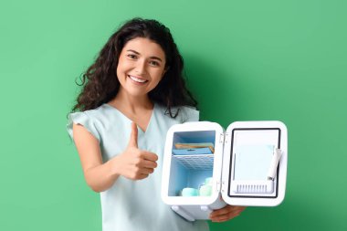 Gülümseyen kadın kozmetik buzdolabını açtı ve yeşil arka planda baş parmağını gösterdi.