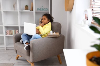 Küçük Afro-Amerikalı kız evdeki koltukta online öğretmenle ders çalışıyor.