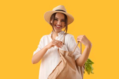 Sarı arka planda eco çantasından şişe alan genç bir kadın.