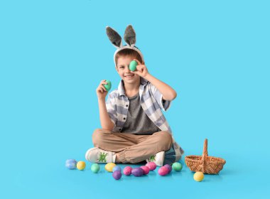 Mavi arka planda tavşan kulaklı ve paskalya yumurtaları olan komik bir çocuk.
