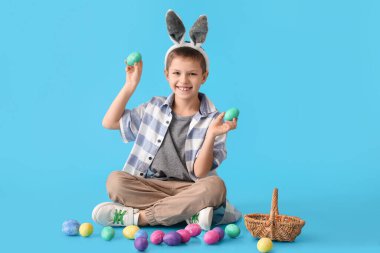 Mavi arka planda tavşan kulaklı ve paskalya yumurtaları olan komik bir çocuk.