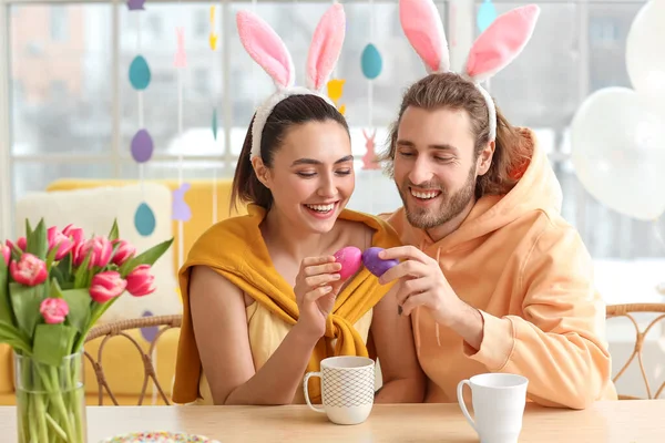 Boldog Pár Nyuszifülekben Húsvéti Tojást Törnek Asztalnál — Stock Fotó