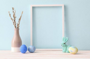 Composition with photo frame, Easter eggs, bunny and pussy willow branches on table near color wall