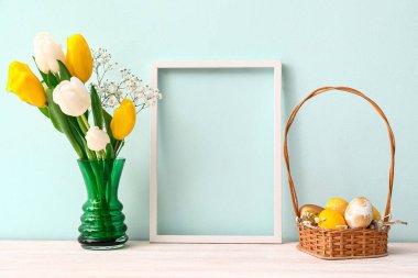 Fotoğraf çerçeveli kompozisyon, sepette Paskalya yumurtası ve renkli duvarın yanındaki masada çiçekler.