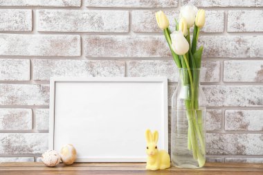 Fotoğraf çerçeveli kompozisyon, Paskalya yumurtaları, tavşan ve çiçekler açık tuğla duvarın yanındaki masada.