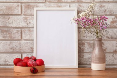 Fotoğraf çerçeveli kompozisyon, Paskalya yumurtaları ve çiçekler açık tuğla duvarın yanındaki masada.