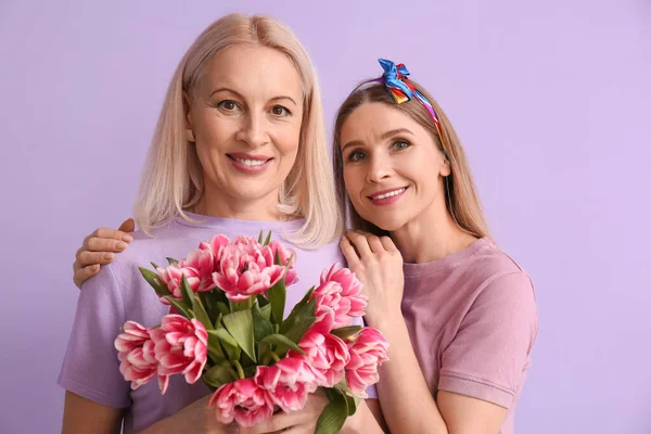 Young woman with her mother and flowers on lilac background