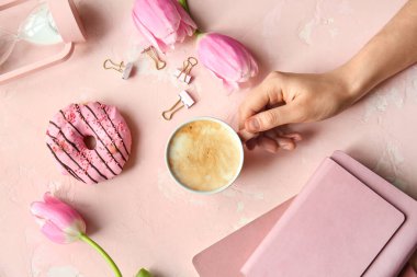 Bir fincan kahve, çörek, not defterleri ve çiçeklerle dolu bir kadın eli.