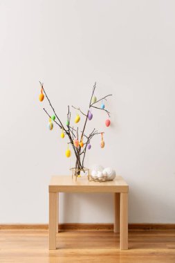 Farklı Paskalya yumurtaları olan ağaç dalları ve aydınlık duvarın yanındaki ahşap masada şık bir dekor.