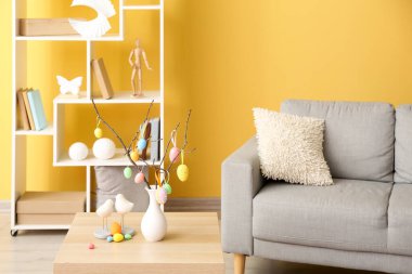 Ağaç dalları ve farklı Paskalya yumurtaları olan vazolar oda içindeki kanepenin yanındaki ahşap masada.