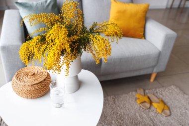 Masanın üzerinde mimoza çiçekleri ve oturma odasının iç tarafında kanepe olan vazo, yakın plan.