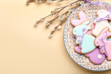 Plate with tasty Easter cookies in shape on bunny and pussy willow branches on beige background, closeup