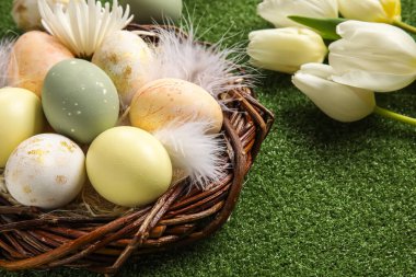 Yeşil arka planda boyanmış Paskalya yumurtaları, tüyleri ve çiçekleri olan yuva, yakın plan.