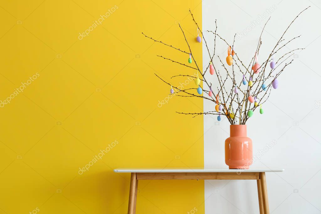 Vase with tree branches and Easter eggs on table near color wall in room