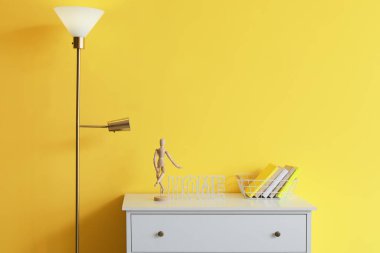 Ahşap mankenli çekmeceler, kitaplar ve sarı duvarın yanında parlayan lamba.