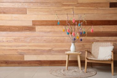 Paskalya kutlaması için süslenmiş ağaç dalları olan vazo masa ve tahta duvarın yanındaki koltukta.