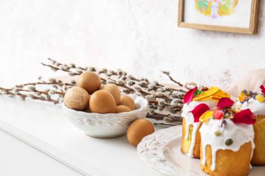 Easter cakes, bowl with eggs and pussy willow branches on chest of drawers near light wall, closeup
