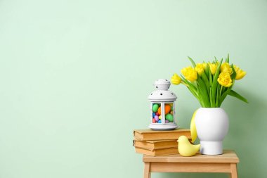 Laleli vazo, kitap yığını, Paskalya yumurtalı fener ve renkli duvarın yanındaki merdivende duran kuş.