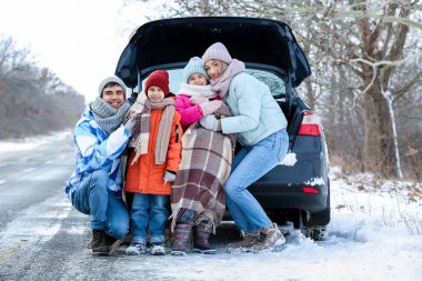 Ebeveynler ve küçük çocukları, karlı kış gününde arabanın yanında kalın giysiler içinde.
