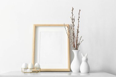 Blank photo frame, holder with Easter eggs and pussy willow branches on table near light wall