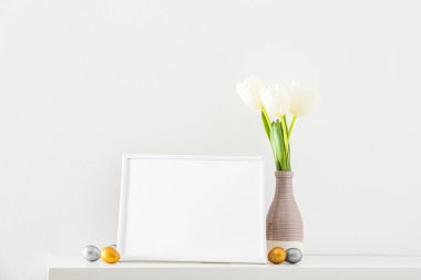 Boş fotoğraf çerçevesi, Paskalya yumurtası ve vazosu lalelerle aydınlık duvarın yanında.