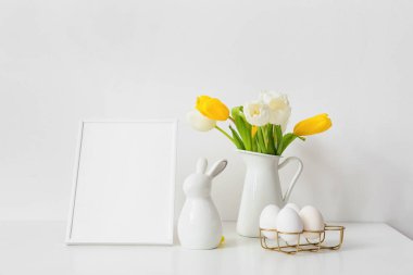 Boş fotoğraf çerçevesi, Paskalya yumurtaları, tavşan ve beyaz duvarın yanındaki masada bir buket lale.